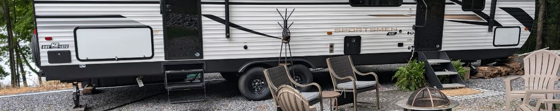 A Sportsmen SE RV parked on gravel with outdoor chairs and a fire pit setup nearby.